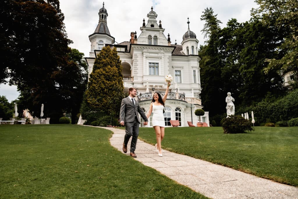 Ein elegantes Paar in förmlicher Kleidung spaziert Hand in Hand auf einem steinernen Weg bei einem großen weißen Herrenhaus in Kärnten, umgeben von üppigem Grün. Hochzeitsfotograf Kärnten