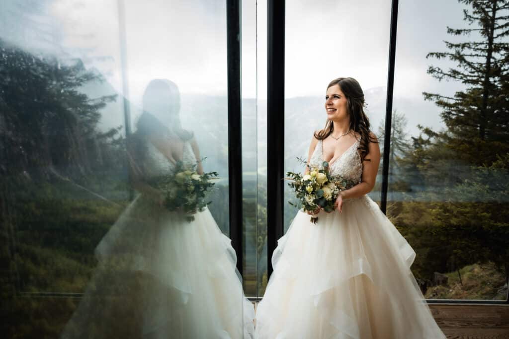 Strahlende Braut im weißen Kleid lächelt durch ein Glasfenster in Kärnten, Spiegelung sichtbar, mit malerischen Bäumen und Bergen im Hintergrund. Hochzeitsfotograf Kärnten