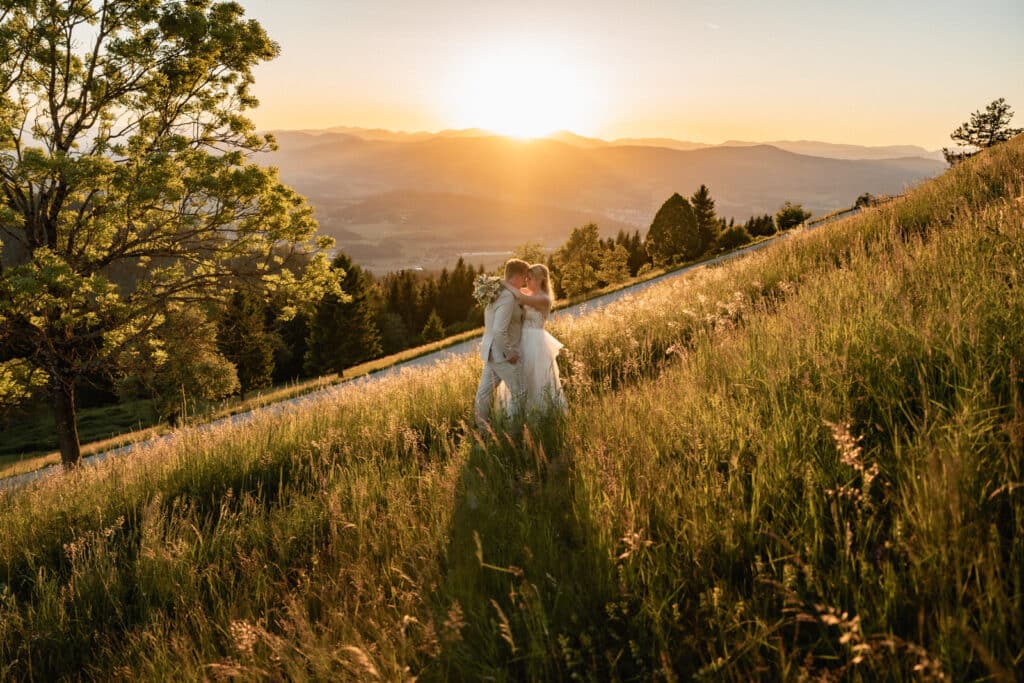 Braut und Bräutigam küssen sich auf einer sonnenbeschienenen Wiese in Kärnten, umrahmt von hohen Bäumen, Bergen und einem goldenen Sonnenuntergang, festgehalten von einem Hochzeitsfotografen. Hochzeitsfotograf Kärnten