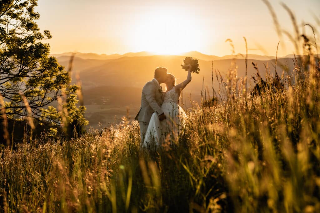 Ein verliebtes Paar steht eng umschlungen in einem goldenen Feld bei weichem Sonnenlicht, sie küssen sich liebevoll und die Braut hält den Brautstrauß in die Luft - eine natürliche und emotionale Momentaufnahme bei einem Outdoor - Fotoshooting