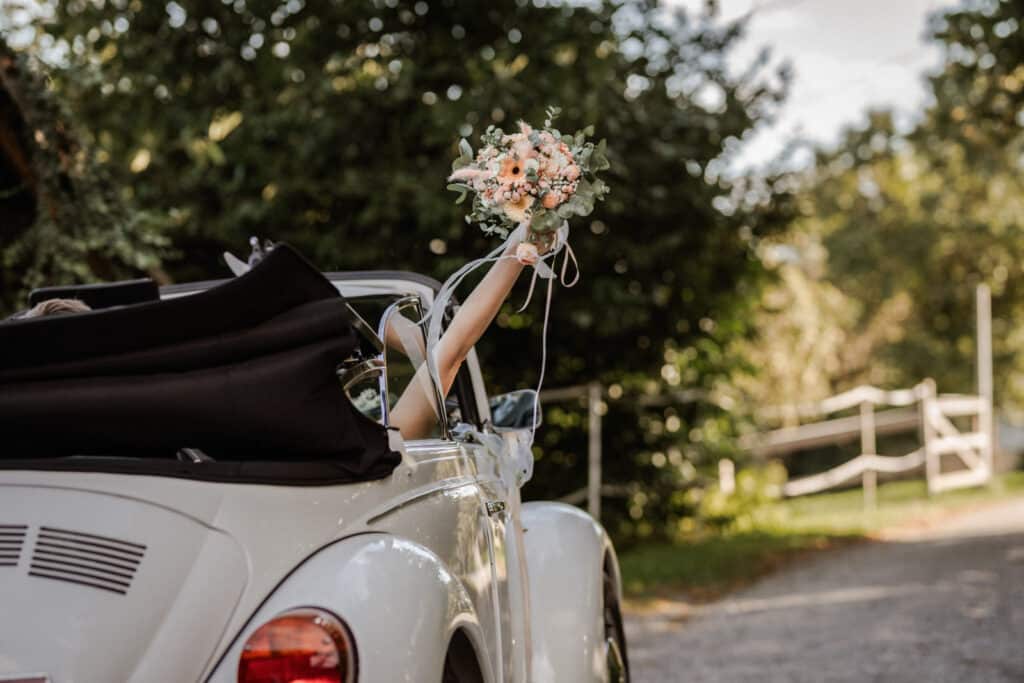 Braut hält Brautstrauß mit Rosen und Eukalyptus aus einem Oldtimer ins Freie