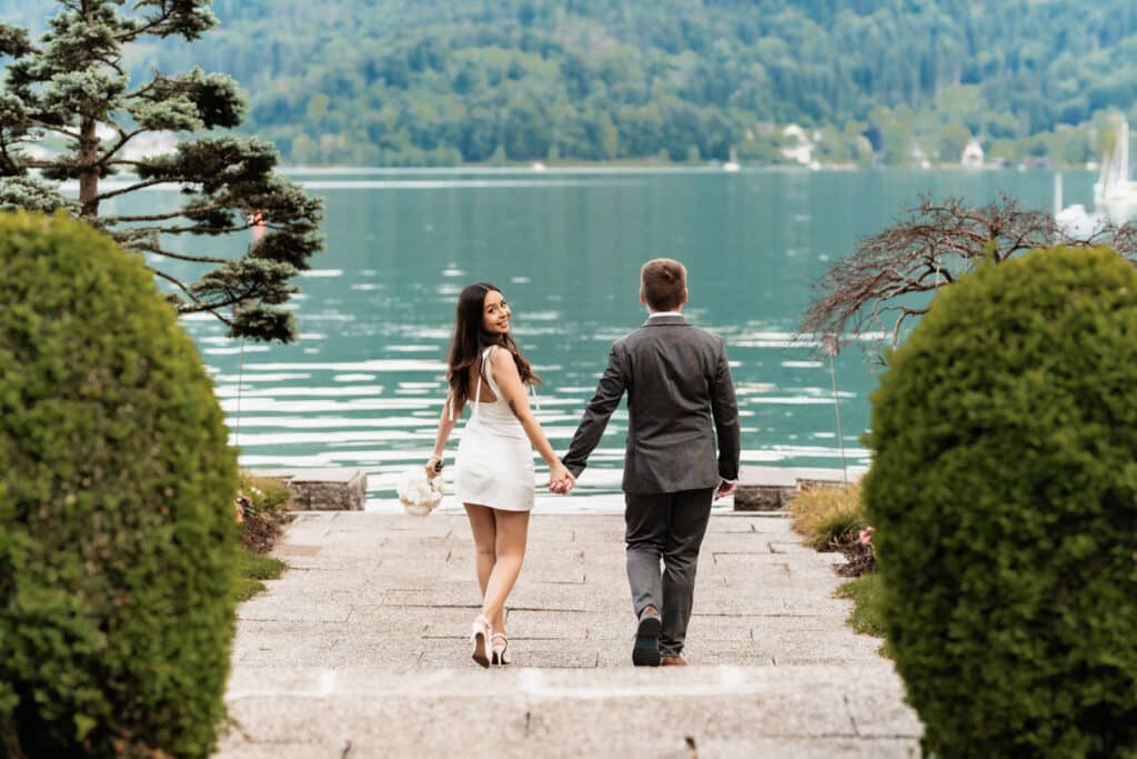 Ein formell gekleidetes Paar geht Hand in Hand zu einem ruhigen See in Kärnten, Österreich, umgeben von üppigem Grün und Bergen. Hochzeitsfotograf Kärnten