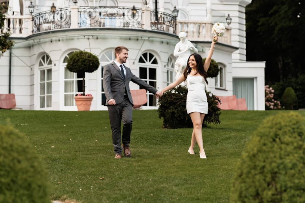 Ein stilvolles Paar spaziert Hand in Hand auf einer Wiese vor einem eleganten weißen Kärntner Gebäude und feiert ausgelassen; Sträucher und Statue sichtbar. Hochzeitsfotograf Kärnten