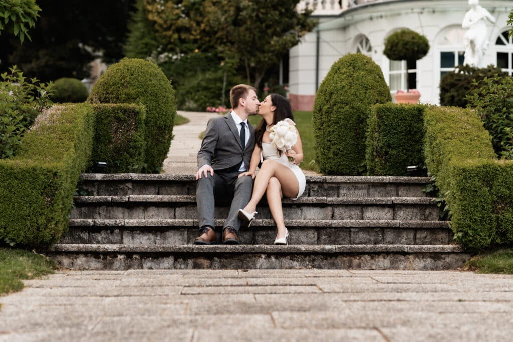 Ein elegantes Paar in formeller Kleidung küsst sich auf einer Steintreppe in Kärnten, mit üppigen Büschen, einem Blumenstrauß und einem großen Gebäude im Hintergrund. Hochzeitsfotograf Kärnten