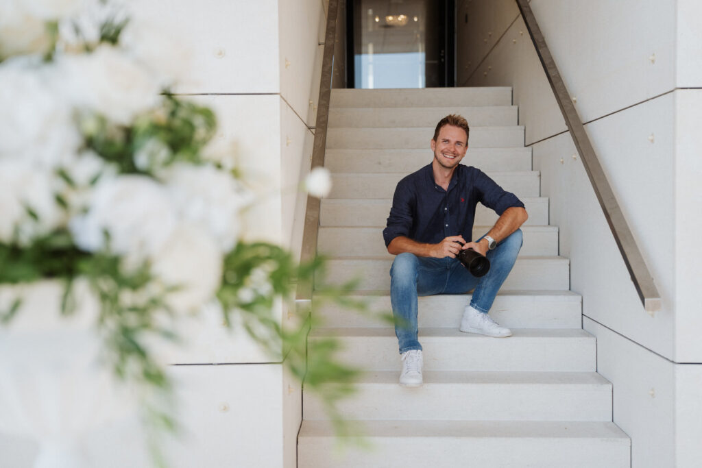 Ein lächelnder Mann sitzt auf einer weißen Treppe und hält eine Kamera. Er trägt ein dunkles Hemd, Jeans und weiße Turnschuhe - perfekt für einen Kontakt Hochzeitsfotograf. Unscharfe weiße Blumen erscheinen im Vordergrund links.